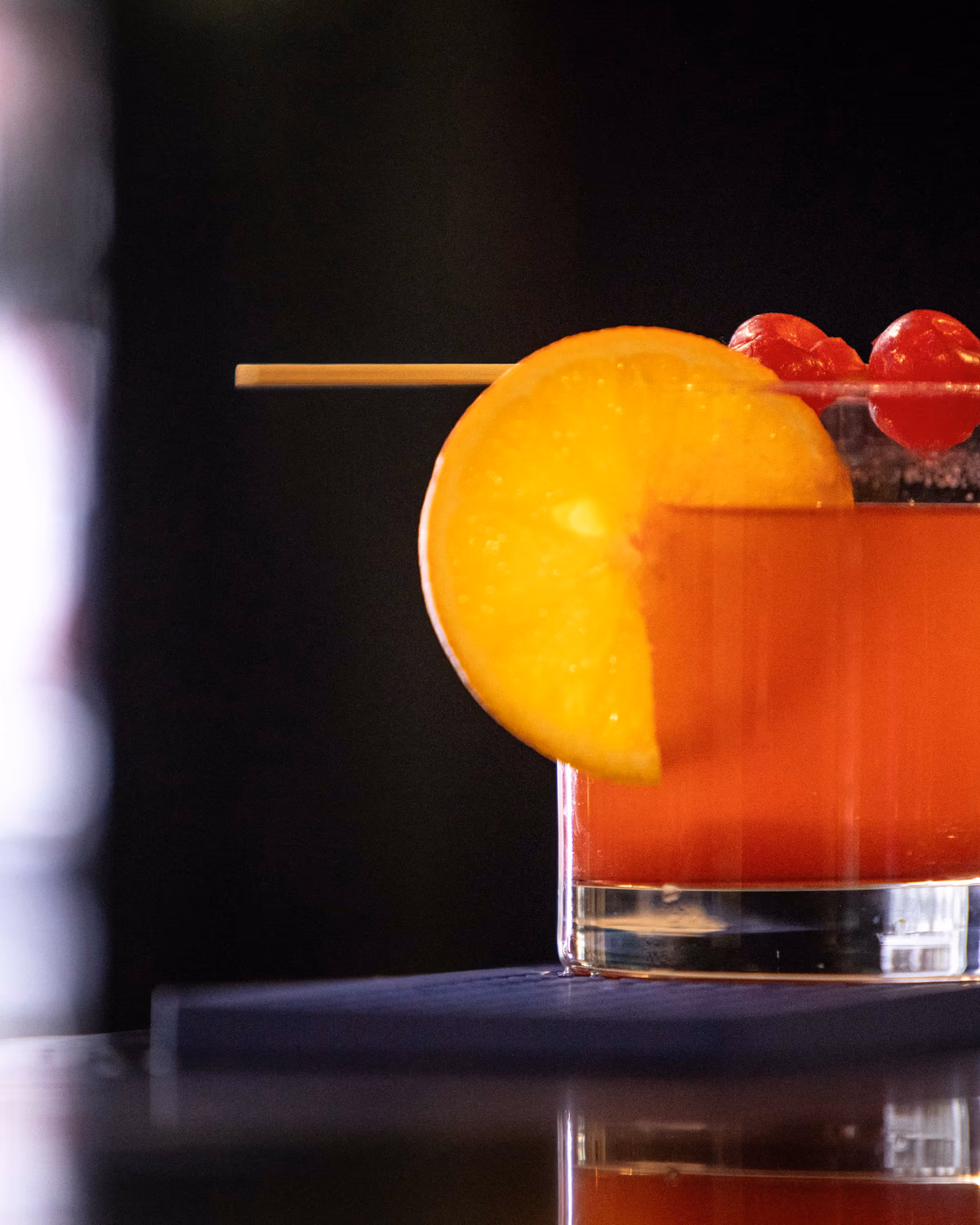 A red signature cocktail garnished with orange and cherries